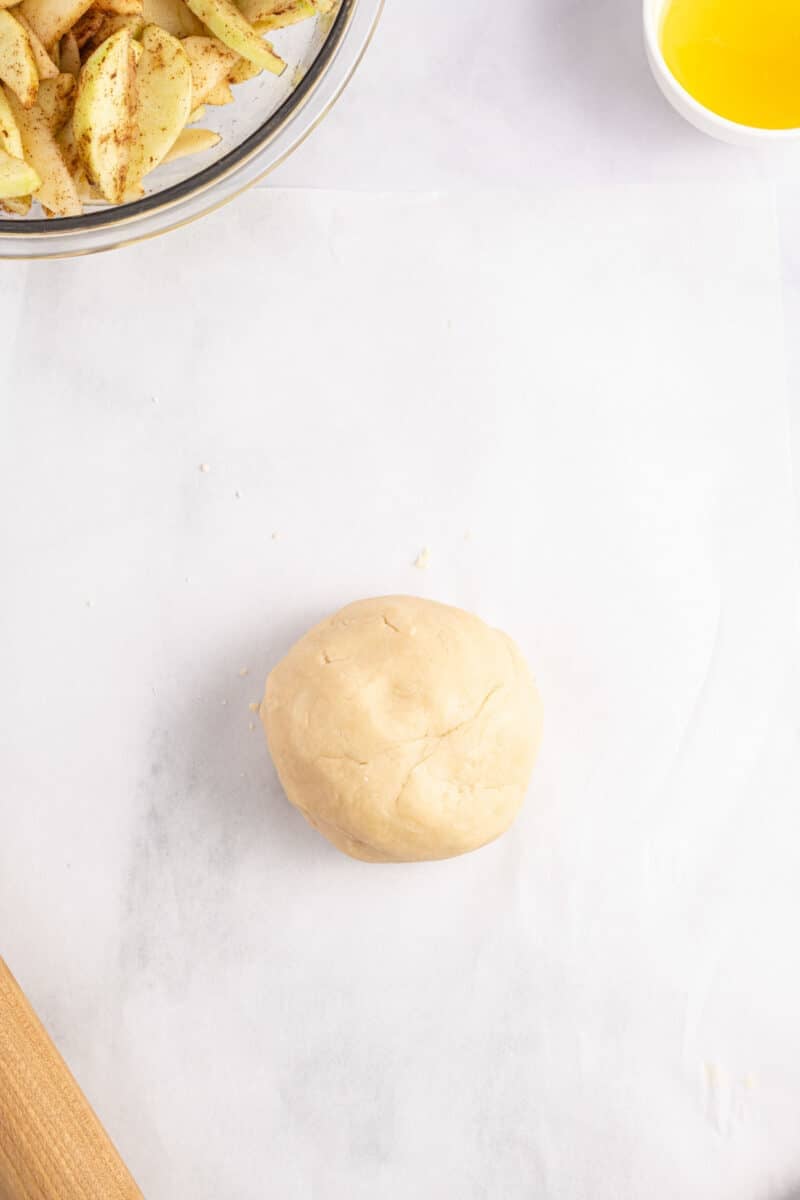 A ball of dough on parchment paper, with a bowl of sliced apples and a dish of melted butter nearby.