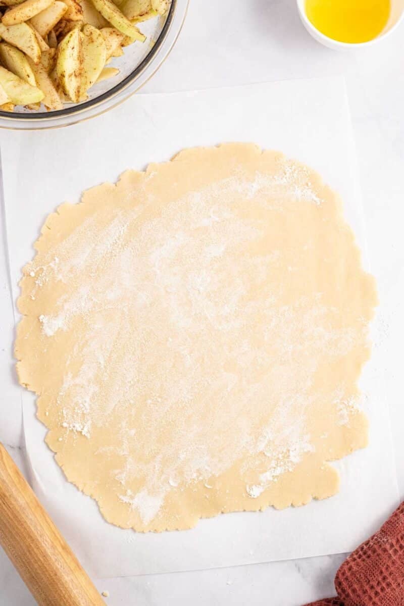 A rolled-out circle of pie dough on parchment paper, with a bowl of sliced apples and a small dish of melted butter nearby.