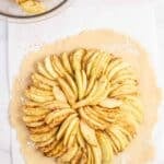Sliced apples arranged in a circular pattern on a round sheet of pie dough, with a bowl of apple slices and a small dish of melted butter nearby.