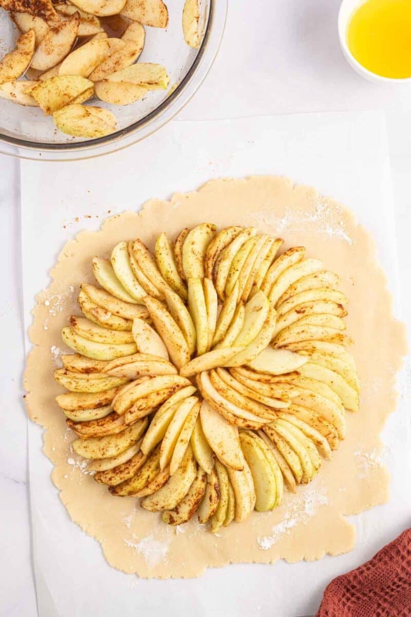 Sliced apples arranged in a circular pattern on a round sheet of pie dough, with a bowl of apple slices and a small dish of melted butter nearby.
