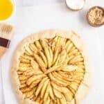 Unbaked rustic apple tart on parchment paper, next to small bowls of butter, sugar, brown sugar, and a pastry brush.