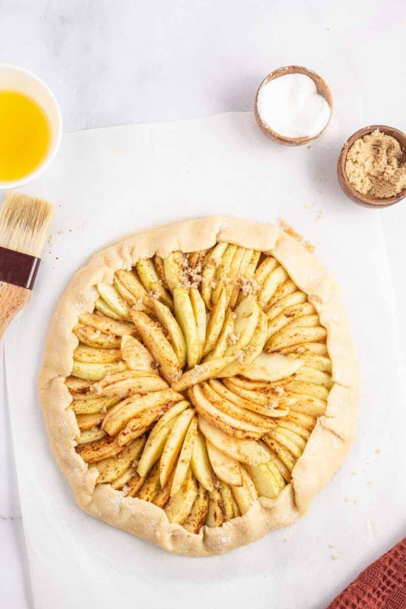 Unbaked rustic apple tart on parchment paper, next to small bowls of butter, sugar, brown sugar, and a pastry brush.