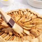 A pastry brush applies glaze to a raw apple galette.