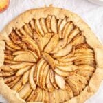 Rustic apple tart with thinly sliced apples arranged in a circular pattern on parchment paper, with whole apples and a plate nearby.