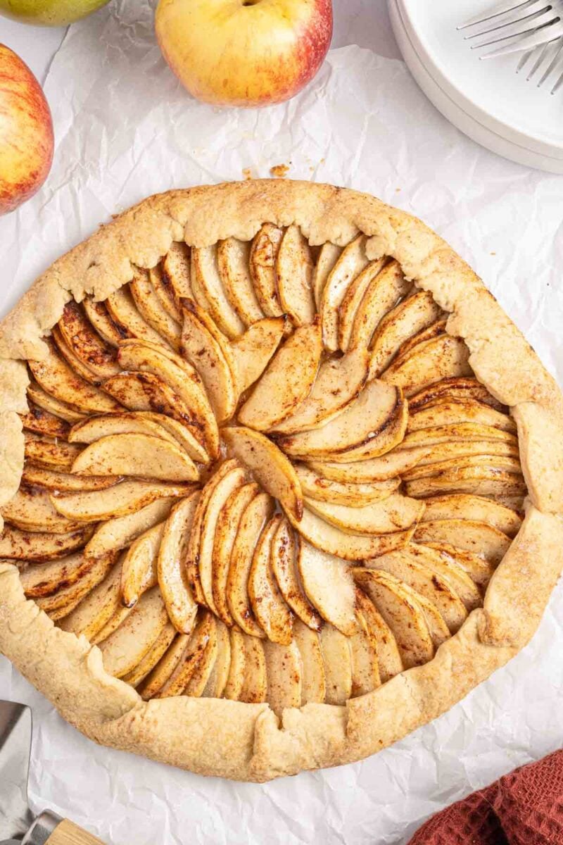 Rustic apple tart with thinly sliced apples arranged in a circular pattern on parchment paper, with whole apples and a plate nearby.