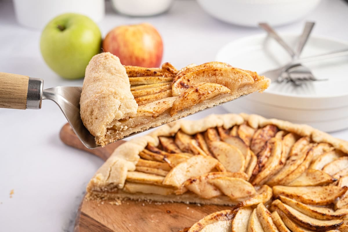 A slice of rustic apple tart being lifted from the whole tart on a metal server.