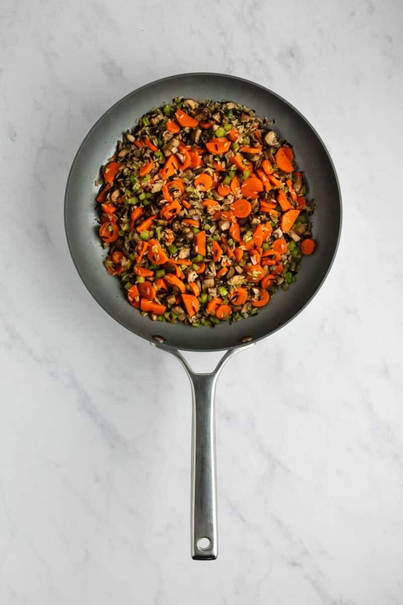 A skillet filled with a sautéed mixture of sliced carrots, celery, mushrooms, and grains.