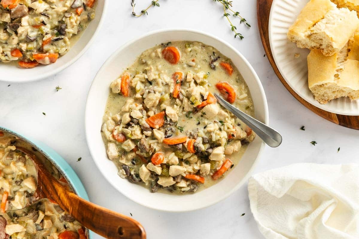 A bowl of chicken and wild rice soup with carrots and mushrooms, served with slices of crusty bread.