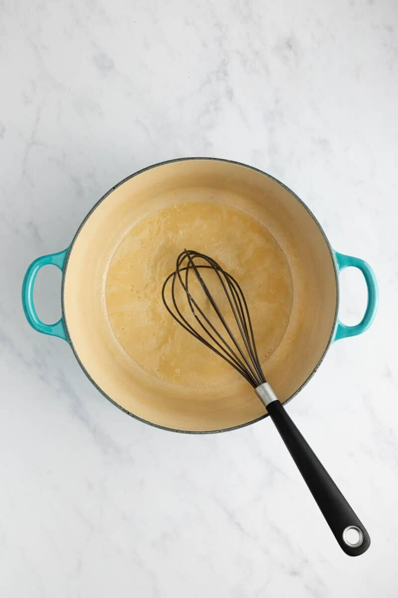 A whisk inside a teal enameled pot containing a light brown liquid base on a marble surface.