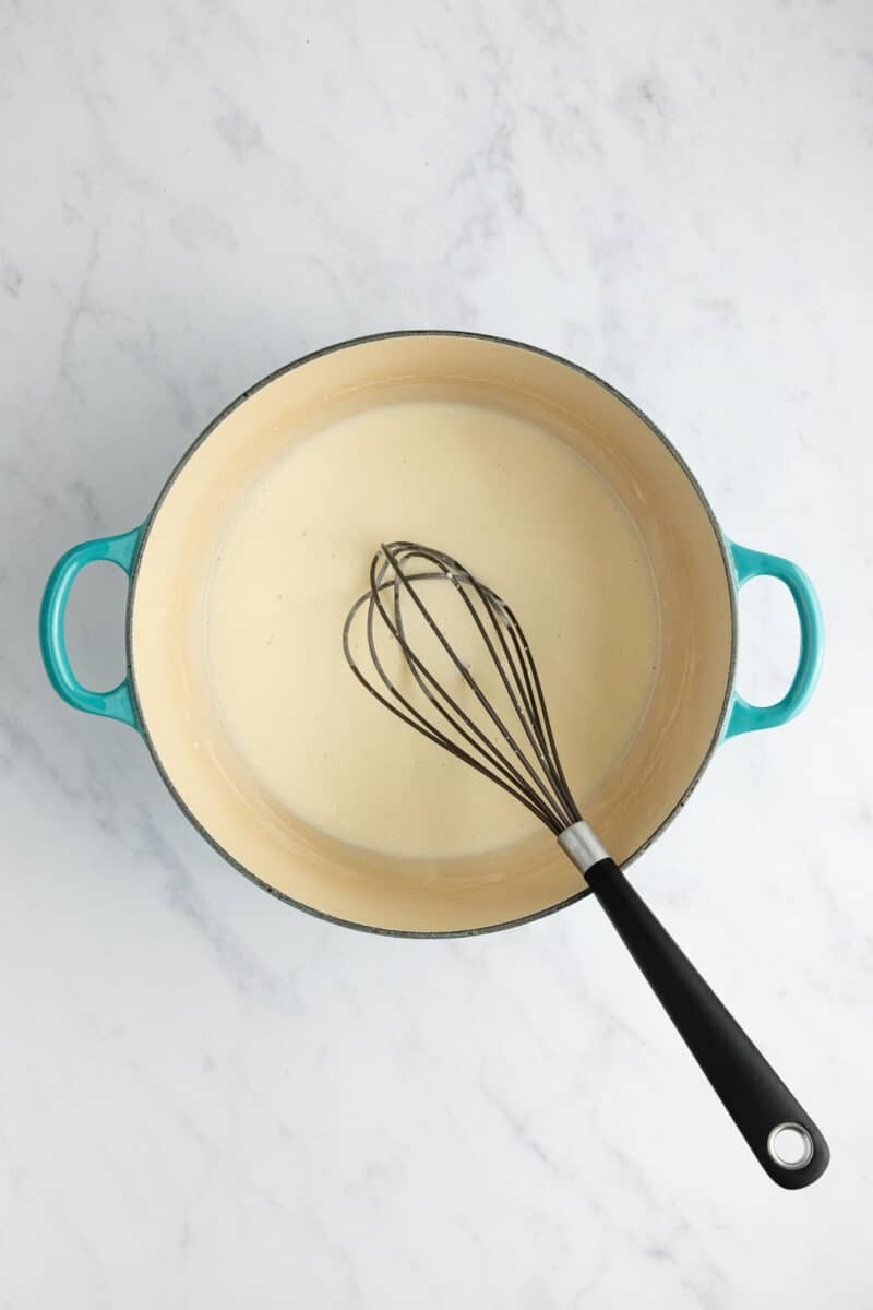 A wire whisk resting in a light-colored sauce inside a teal pot.