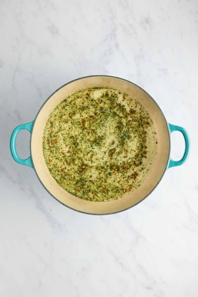 A turquoise enameled Dutch oven filled with a light-colored liquid seasoned with green herbs and red pepper flakes.