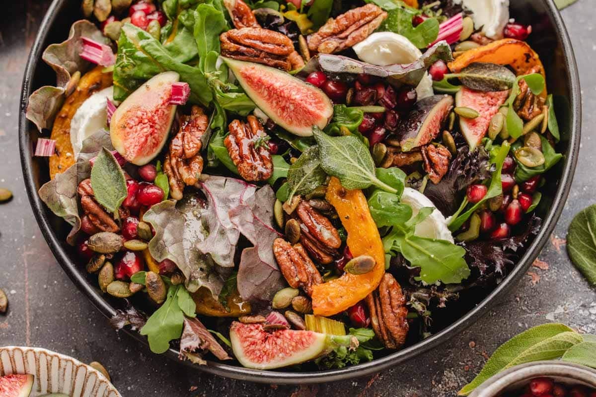 A salad in a dark bowl featuring fresh figs, pecans, pomegranate seeds, roasted squash, and leafy greens.