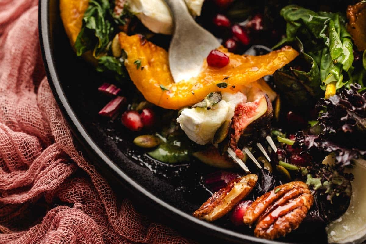A fork lifting a piece of roasted squash, fresh fig, and goat cheese from a salad with pecans and pomegranate seeds.