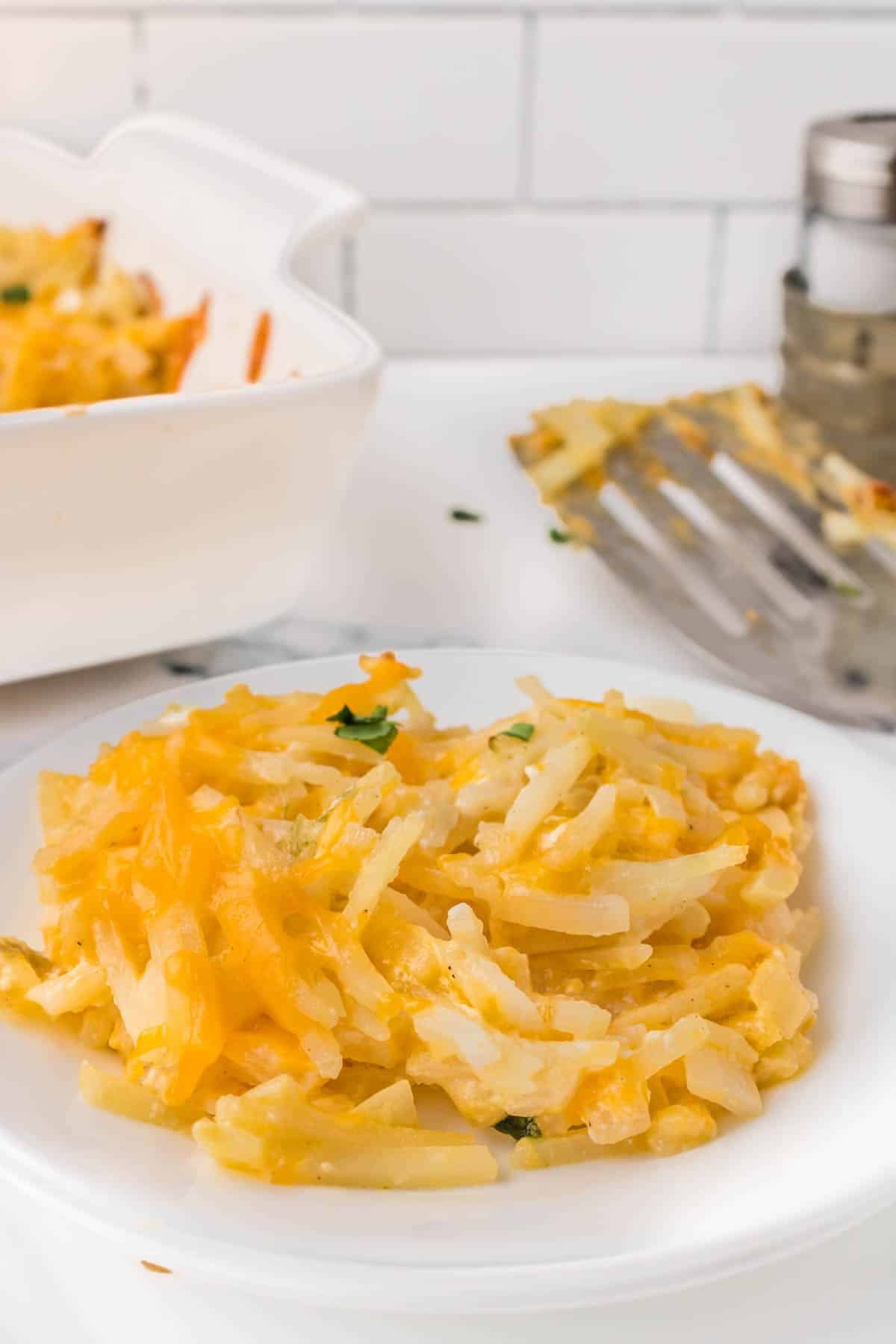 A serving of cheesy shredded potatoes on a white plate, with a baking dish and spatula in the background.
