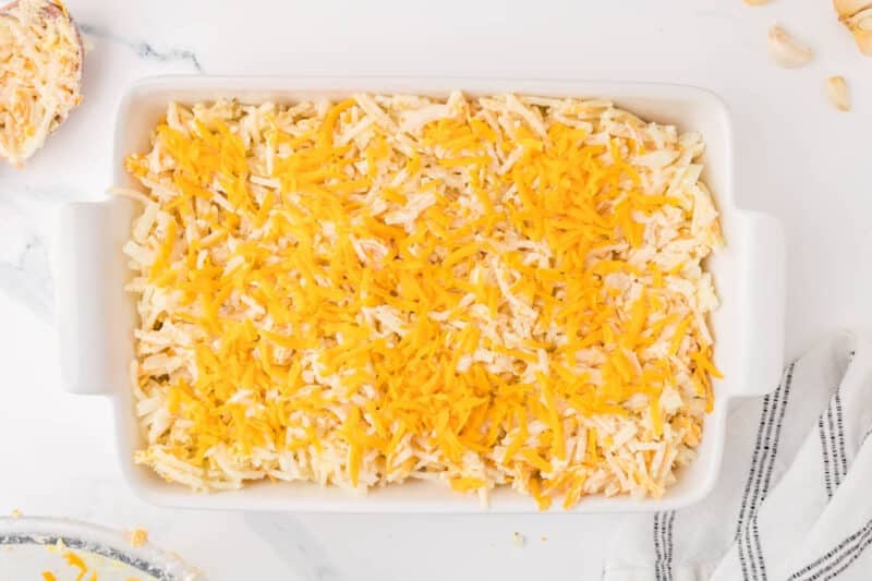 A rectangular white baking dish filled with a mixture of shredded potatoes and topped with shredded cheddar cheese.