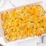A rectangular white baking dish filled with cheesy hash brown casserole topped with chopped green herbs.