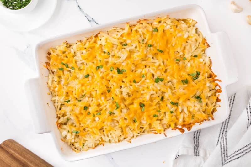 A rectangular white baking dish filled with cheesy hash brown casserole topped with chopped green herbs.
