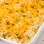 Hash brown casserole topped with melted shredded cheddar cheese and green herbs in a white baking dish.