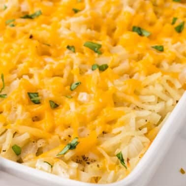 Hash brown casserole topped with melted shredded cheddar cheese and green herbs in a white baking dish.