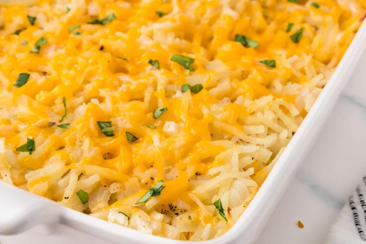 Hash brown casserole topped with melted shredded cheddar cheese and green herbs in a white baking dish.