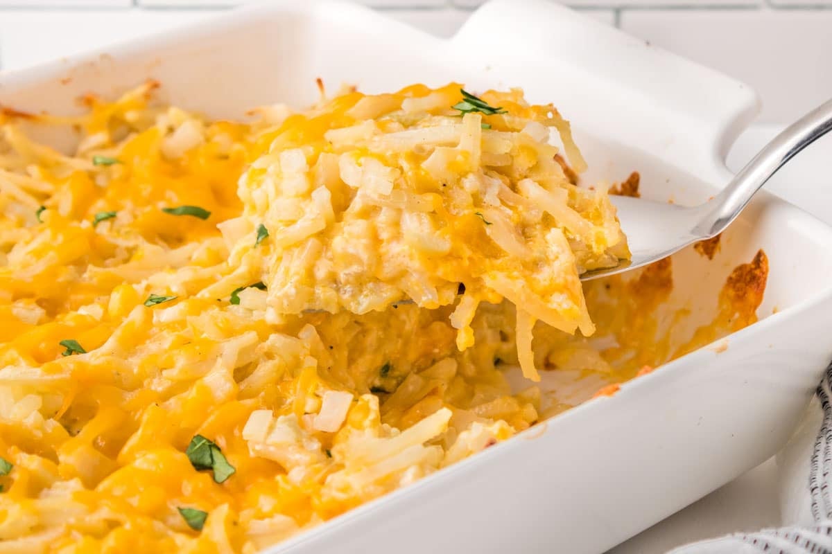 A metal spatula lifts a scoop of cheesy hash brown casserole from a white baking dish.