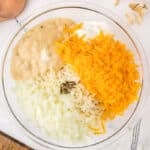 A clear glass bowl filled with chopped onions, shredded cheddar cheese, a dollop of sour cream, creamy soup, and black pepper.