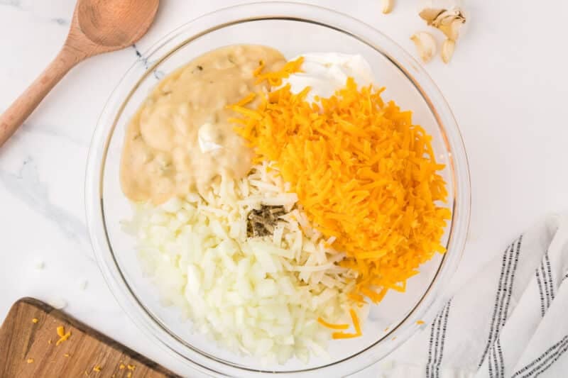 A clear glass bowl filled with chopped onions, shredded cheddar cheese, a dollop of sour cream, creamy soup, and black pepper.