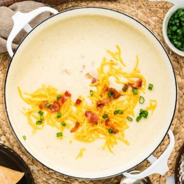 Creamy soup in a white pot topped with shredded cheddar cheese, bacon bits, and chopped green onions.