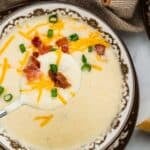 A spoonful of creamy potato soup topped with shredded cheddar, bacon, and green onions, held over a matching bowl of the same soup.
