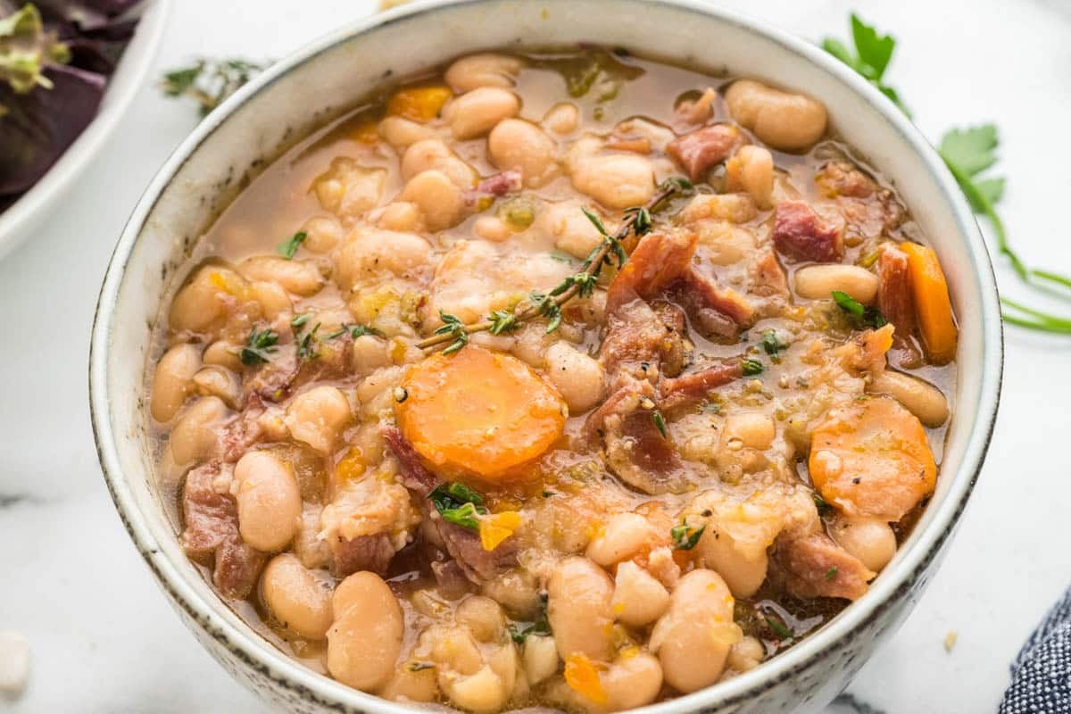 A bowl of navy bean and ham soup with sliced carrots and fresh herbs.