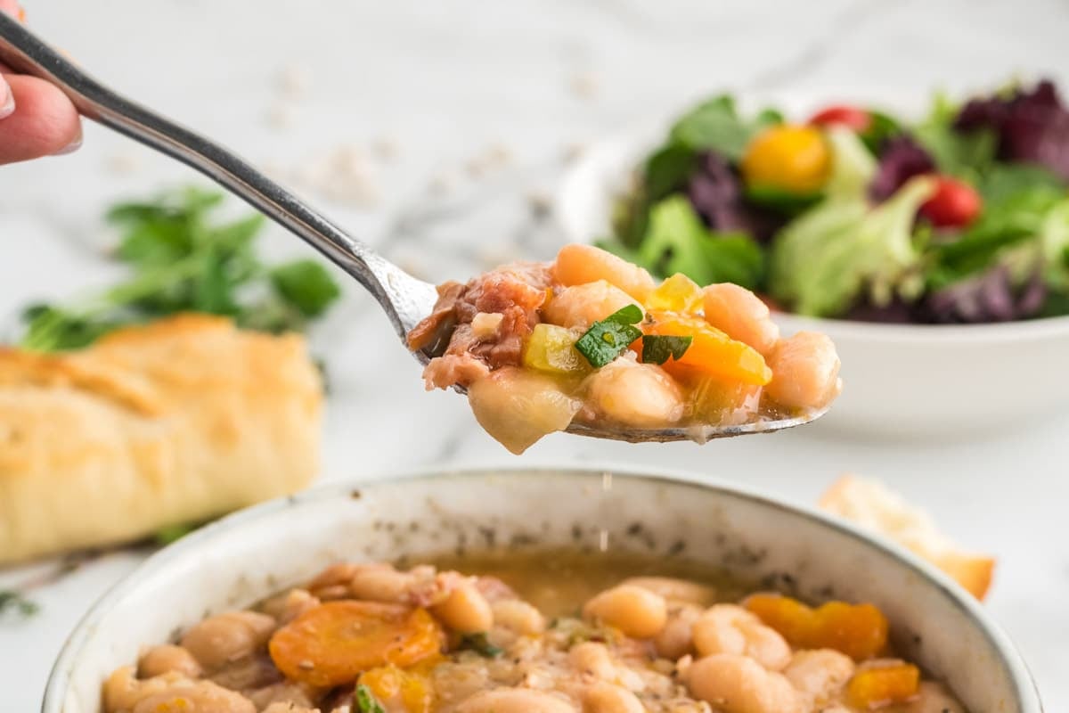 A spoonful of navy bean and ham soup held over a bowl, with a side salad and bread in the background.