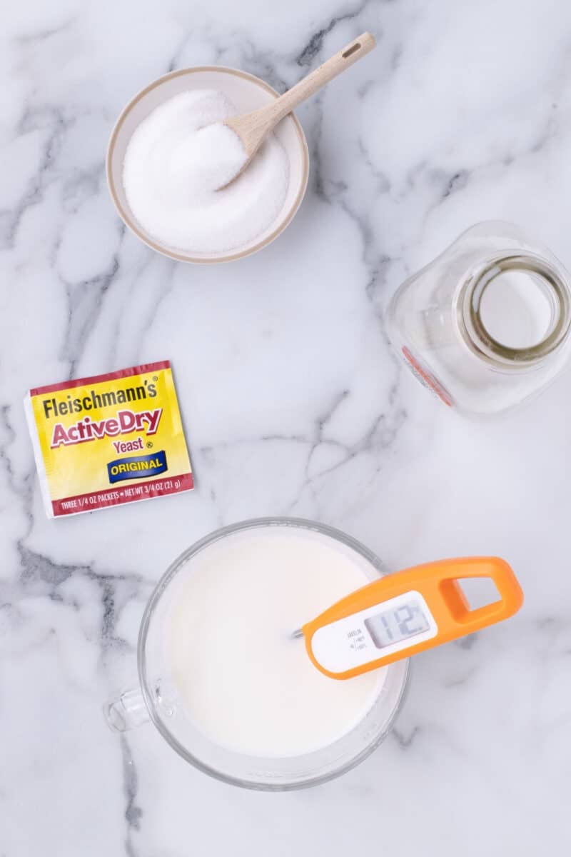 A packet of Fleischmann’s Active Dry Yeast, a small bowl of sugar with a spoon, and a glass measuring cup of milk with an orange digital thermometer reading 112 degrees.
