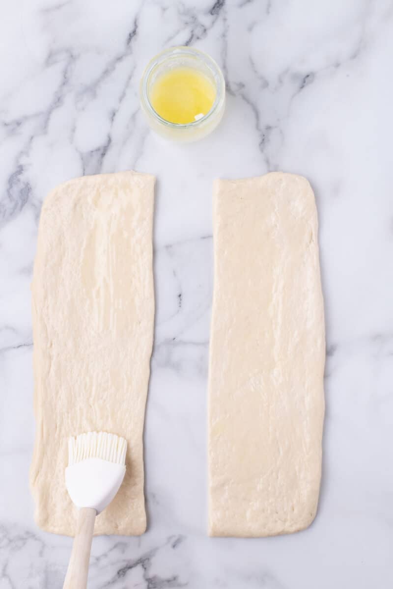 A pastry brush spreading melted butter onto two rectangular pieces of dough on a marble surface.