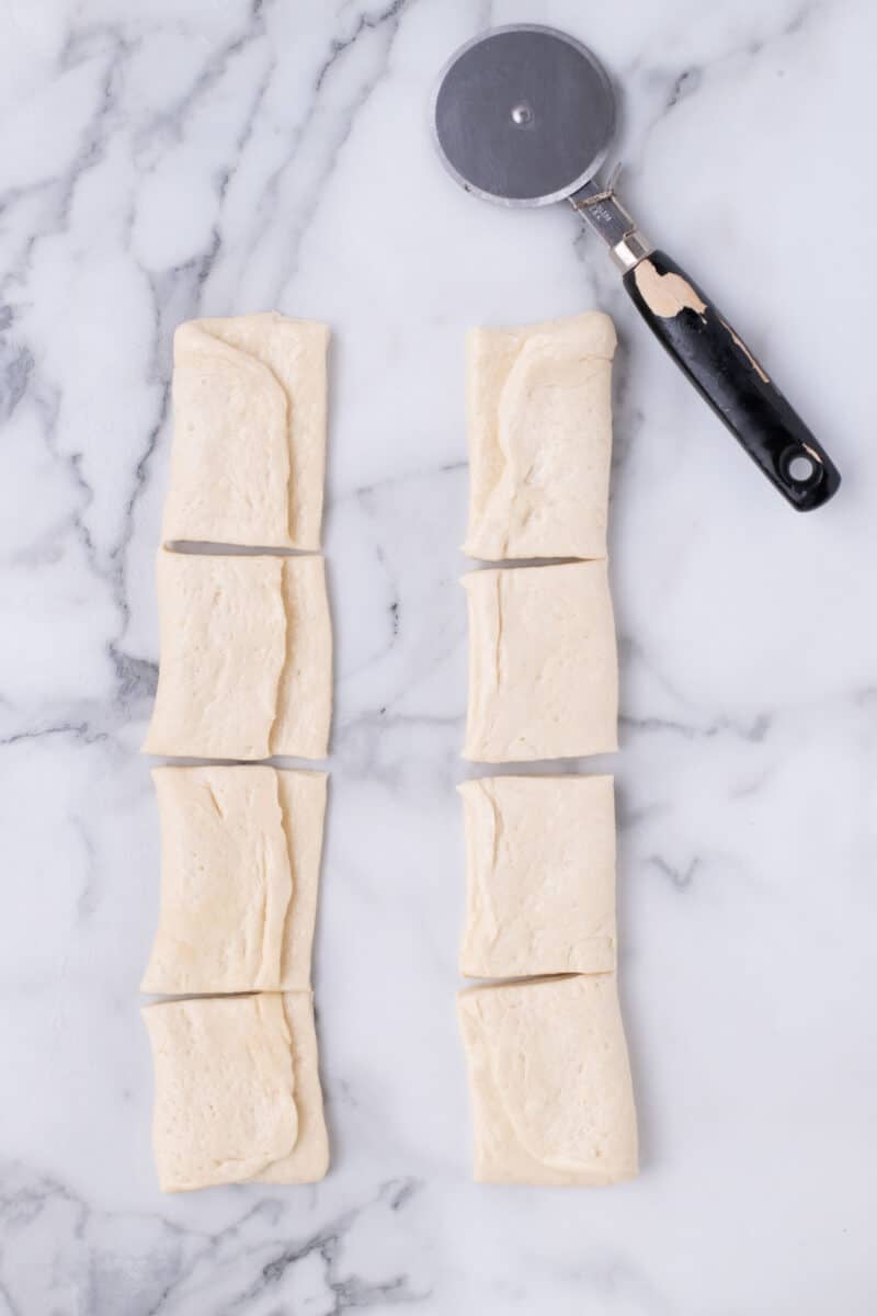 Eight pieces of raw folded dough arranged in two columns on a marble surface next to a pizza cutter.