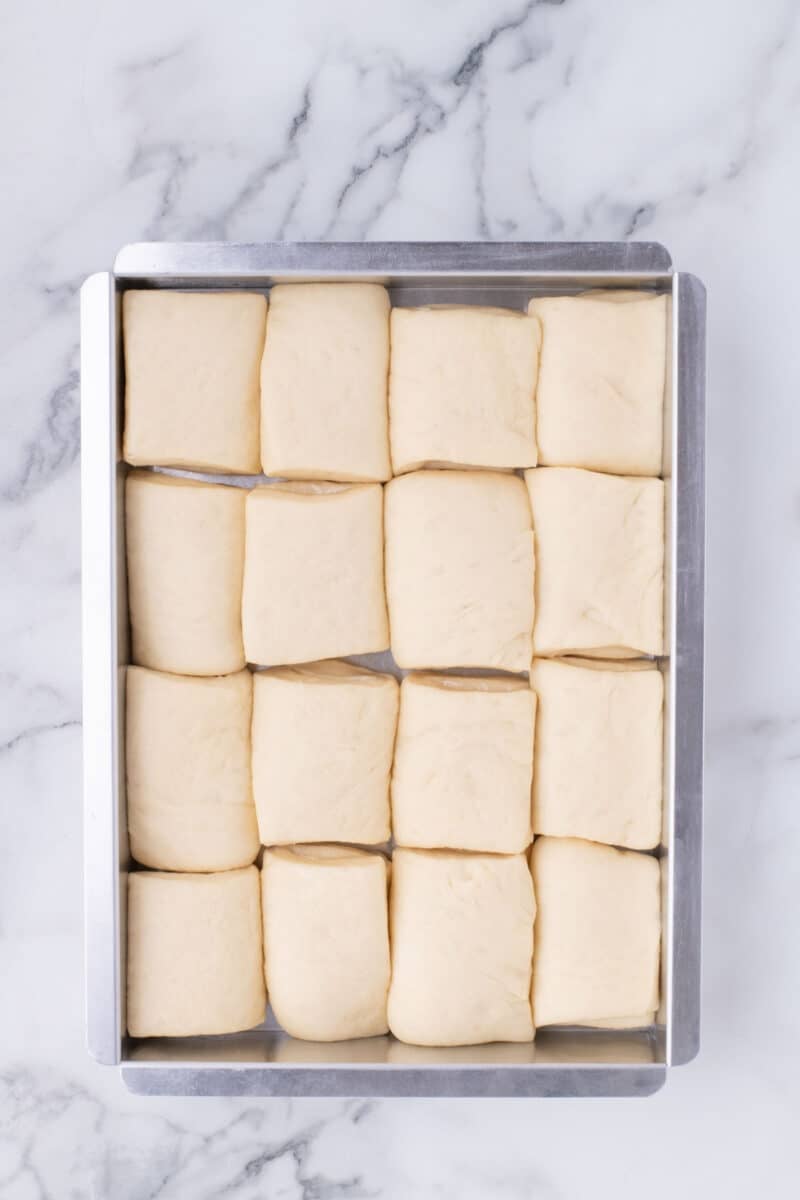 Rectangular metal baking pan filled with sixteen uniform, unbaked dough rolls arranged in a four-by-four grid.
