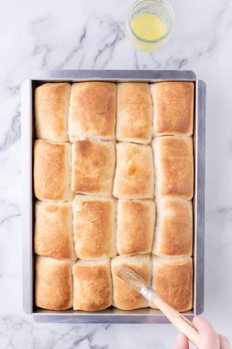 A baking pan filled with golden-brown dinner rolls, with a pastry brush applying butter to one roll and a small bowl of melted butter nearby.