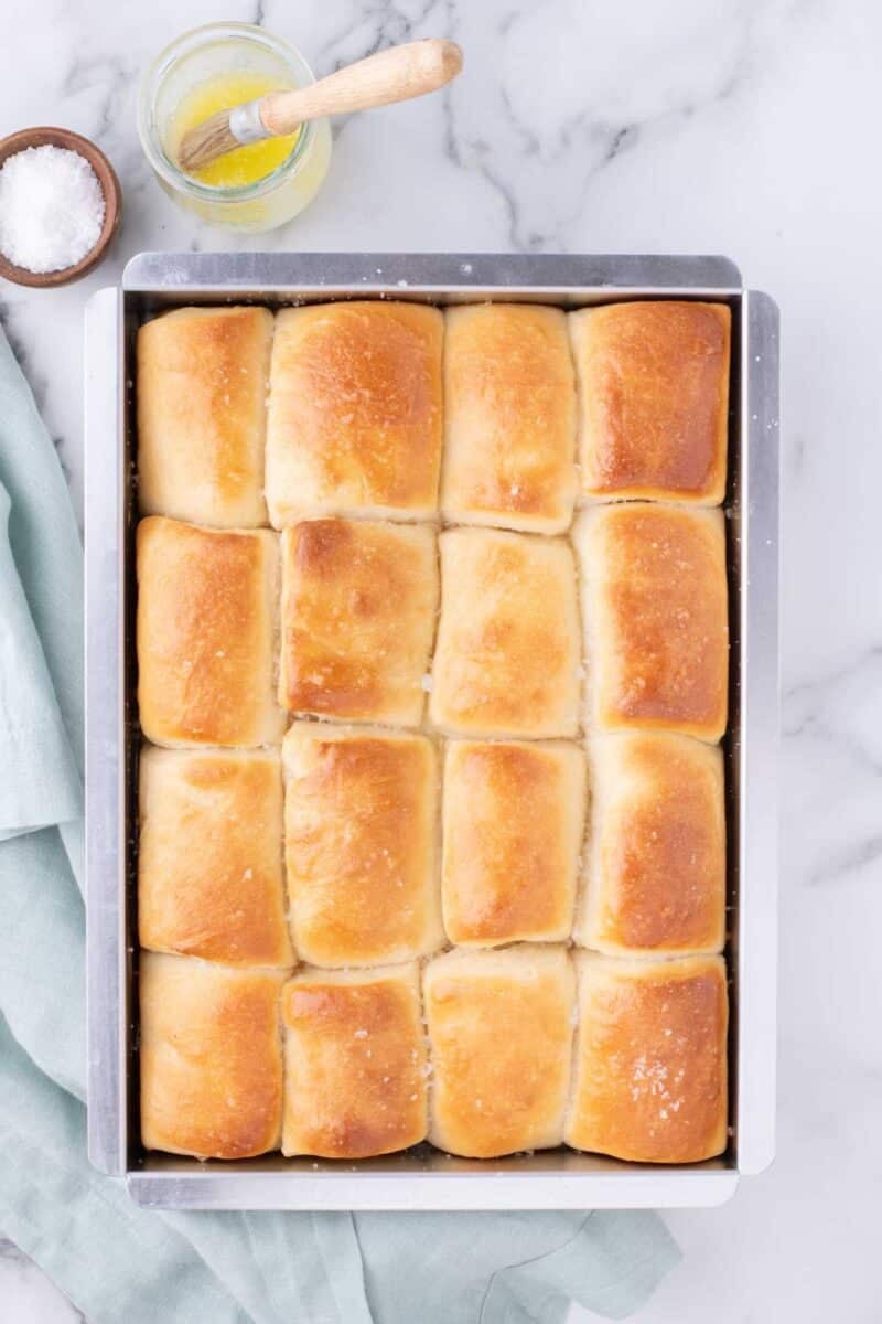 A rectangular pan of sixteen golden-brown dinner rolls, with a small jar of melted butter and a dish of coarse salt nearby.