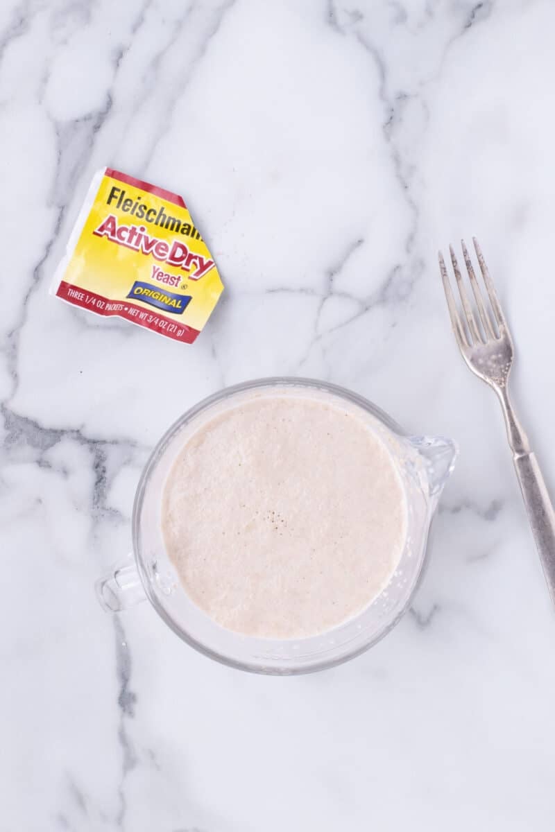 A packet of Fleischmann’s Active Dry Yeast next to a glass measuring cup containing a frothy yeast mixture and a fork on a marble surface.