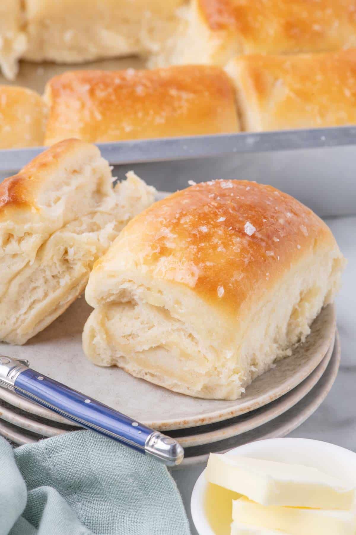 Two golden-brown dinner rolls sprinkled with flaky sea salt on a plate, with a blue butter knife and a dish of butter nearby.