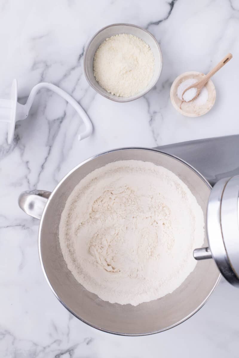 Stand mixer bowl filled with flour, with a dough hook attachment, a bowl of grated cheese, and a small spoonful of salt on a marble countertop.