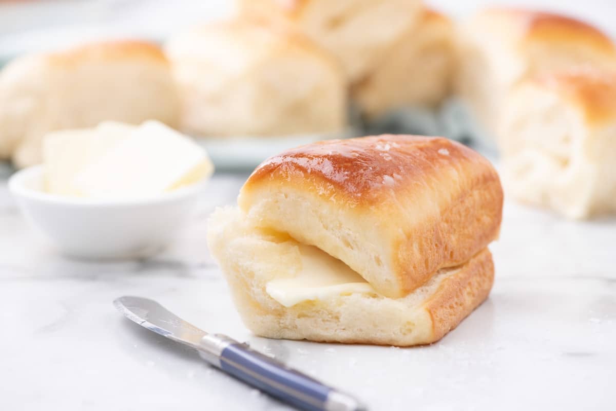 A split dinner roll with butter inside, sitting on a marble surface next to a butter knife and a small bowl of butter pats, with additional rolls in the background.