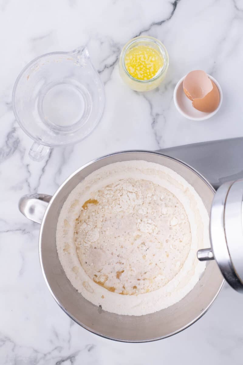 A stand mixer bowl containing flour and liquid ingredients, with a glass measuring cup, melted butter, and an eggshell nearby on a marble surface.