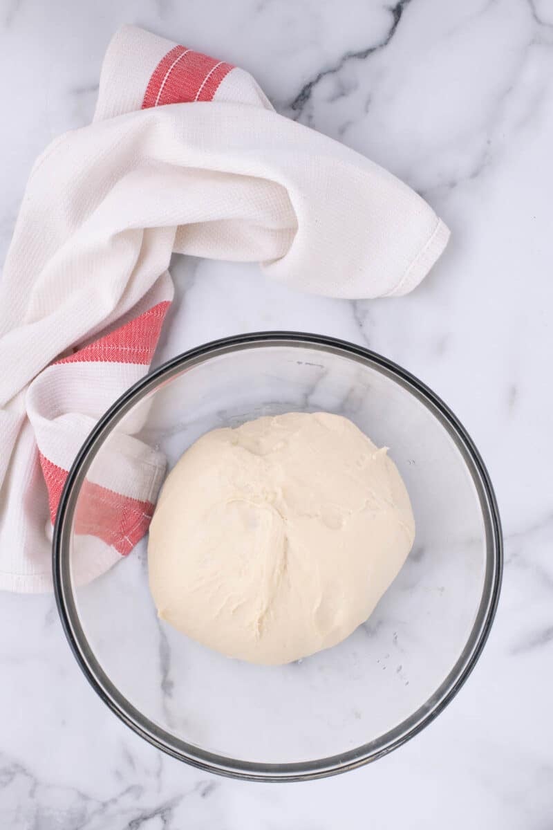 A ball of dough in a clear glass bowl next to a red and white striped kitchen towel on a marble surface.