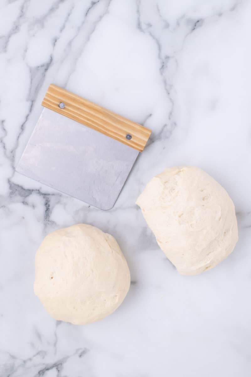 Two balls of dough and a metal bench scraper with a wooden handle on a marble surface.