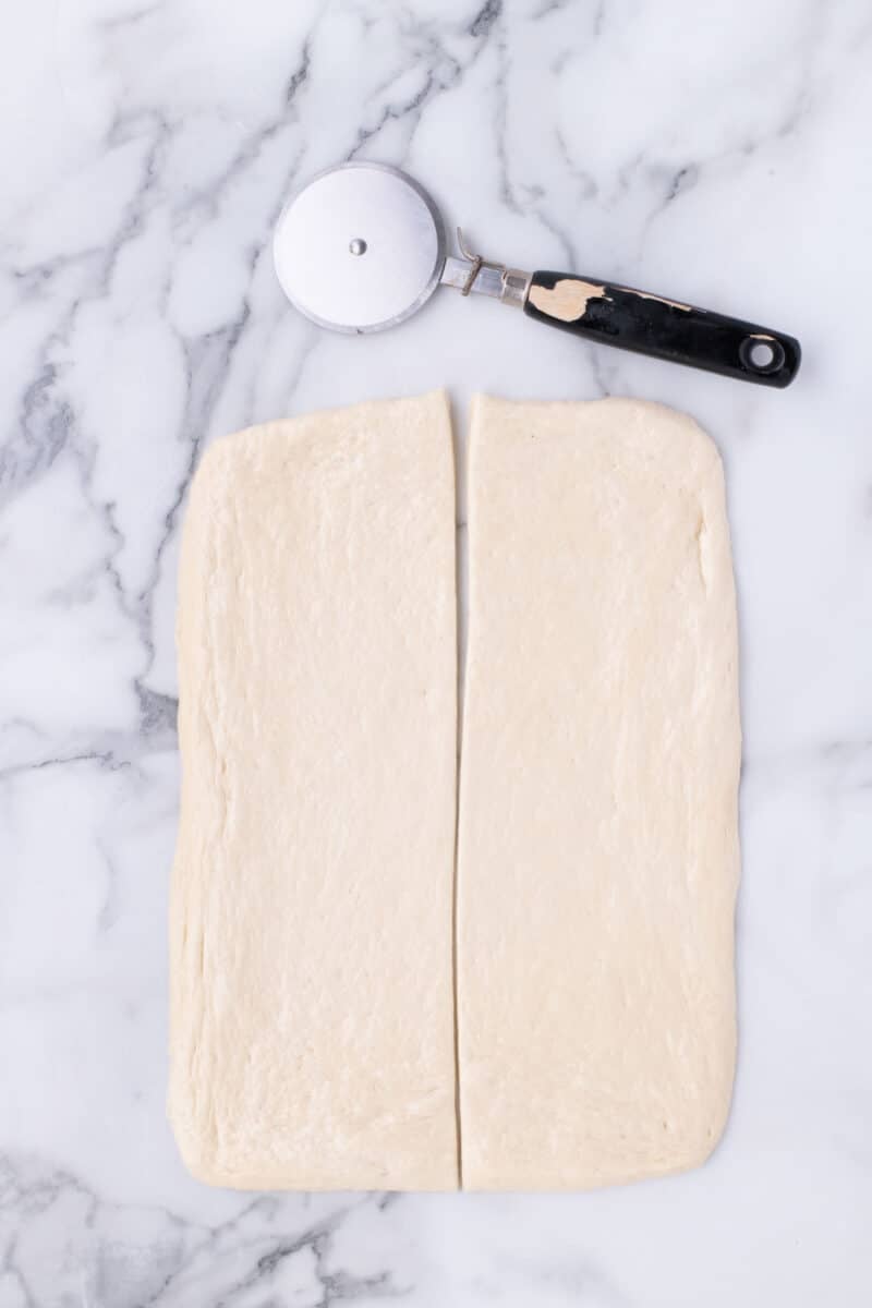 A rectangle of dough sliced into two pieces, with a pizza cutter resting above it.