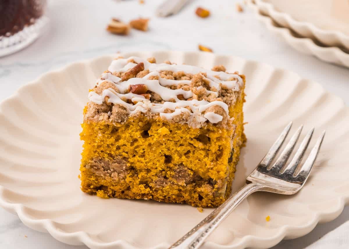 A square of pumpkin coffee cake with a streusel topping and white icing drizzle on a scalloped plate with a silver fork.