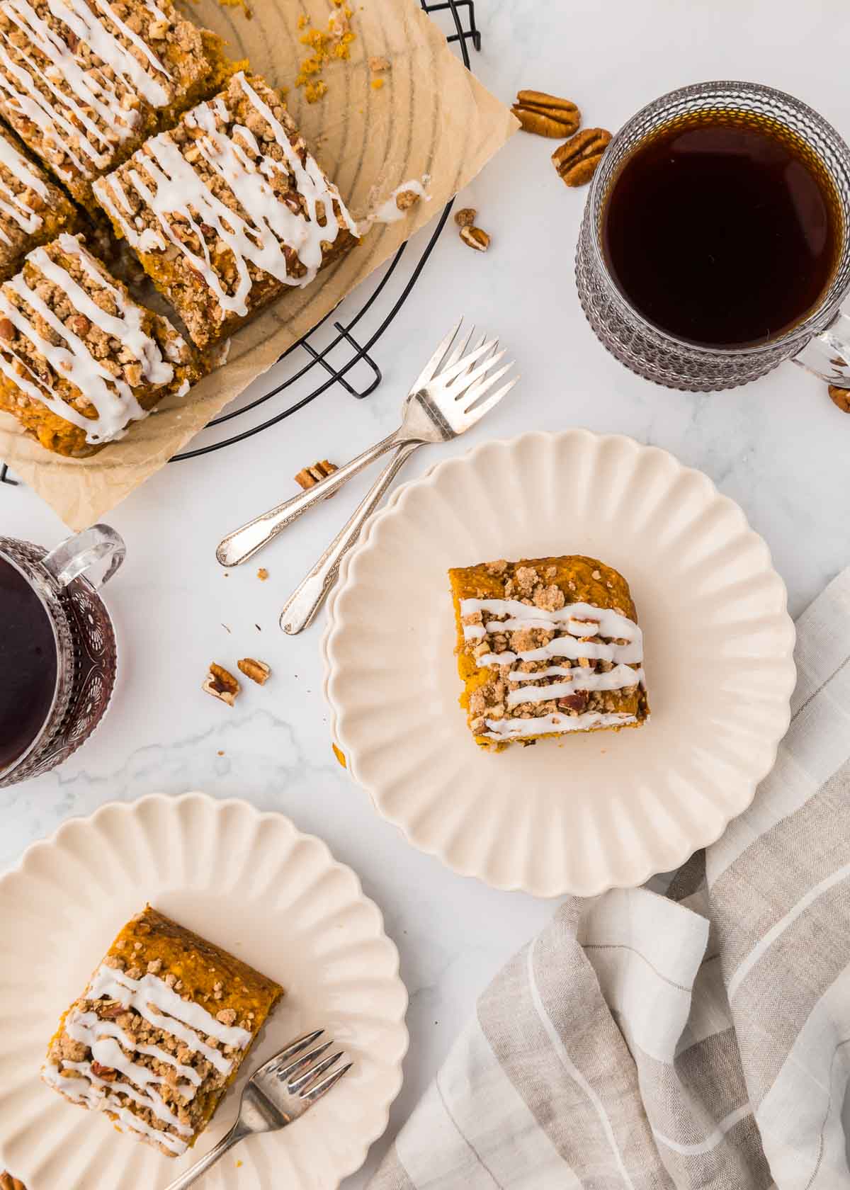 A slice of pumpkin coffee cake with streusel topping and icing on a scalloped plate, next to a larger batch on a wire rack and two cups of coffee.