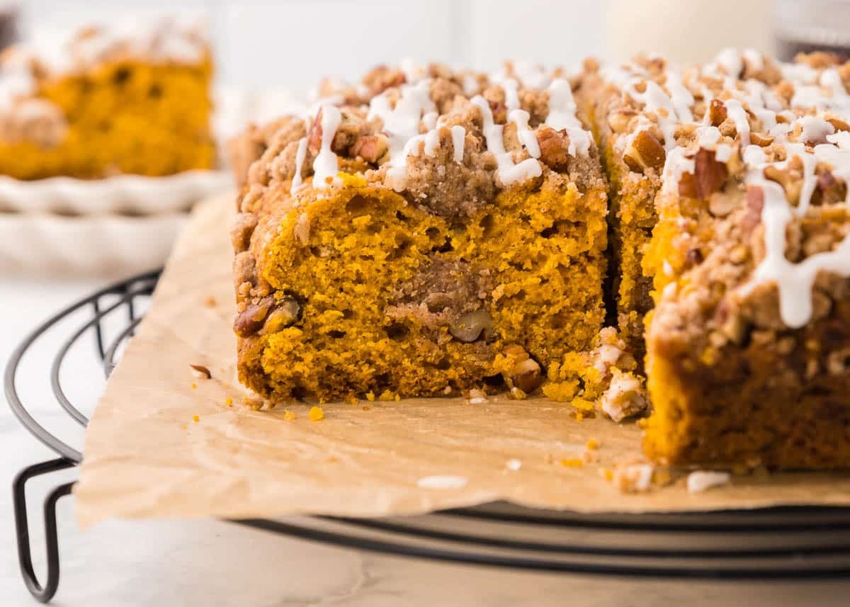 A slice cut from a pumpkin coffee cake topped with nuts and white icing on a wire cooling rack.
