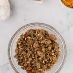 A glass bowl filled with a pecan crumb topping, with bowls of flour, eggs, and pumpkin puree nearby on a white surface.