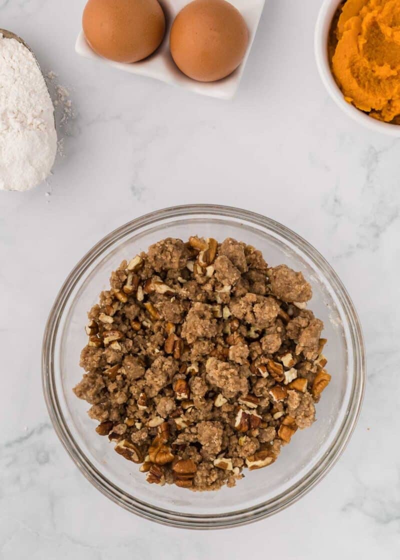 A glass bowl filled with a pecan crumb topping, with bowls of flour, eggs, and pumpkin puree nearby on a white surface.